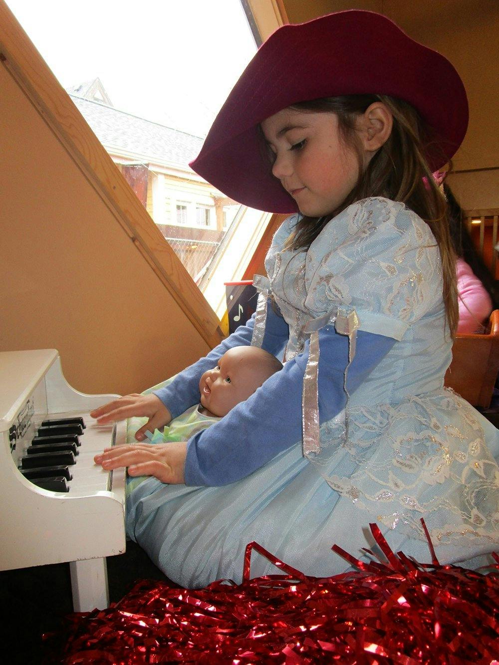 Child in a hat and dress playing a toy piano with a doll