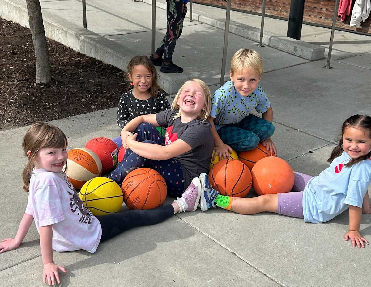 Five children sitting on the ground surrounded by various sports balls