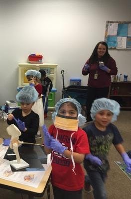 Children wearing surgical caps, masks, and gloves in a classroom