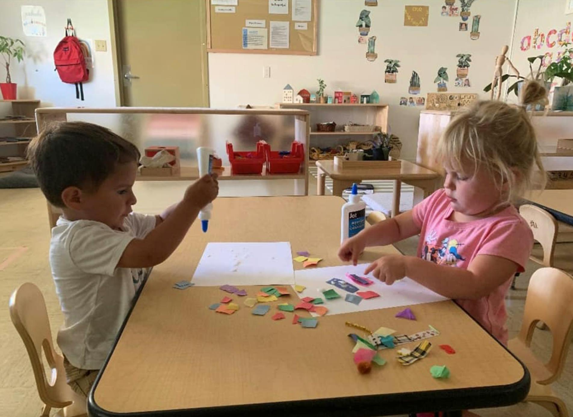 Two children at a table doing a craft with paper scraps and glue