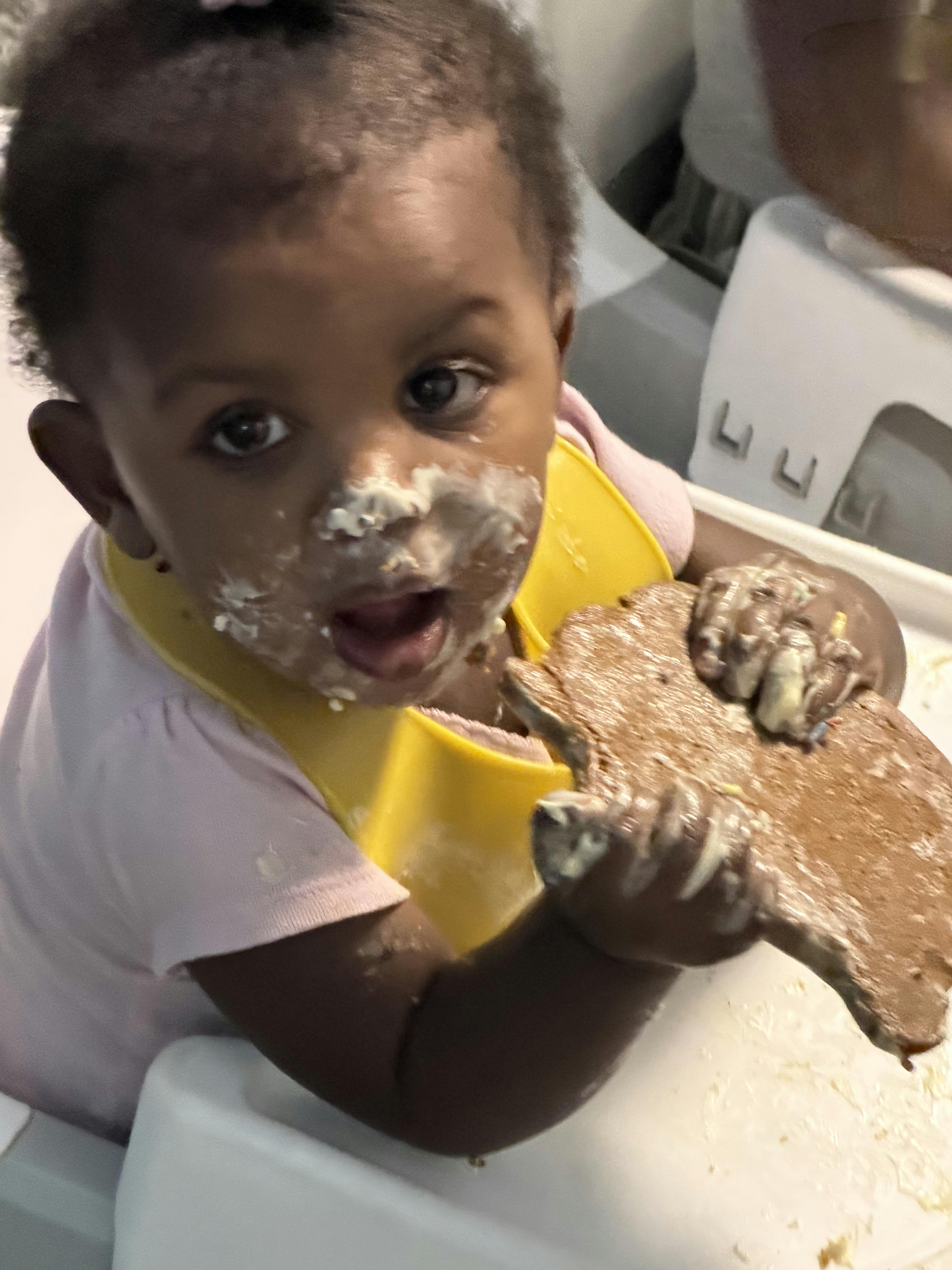 Child in a yellow bib with food on their face, holding a piece of bread