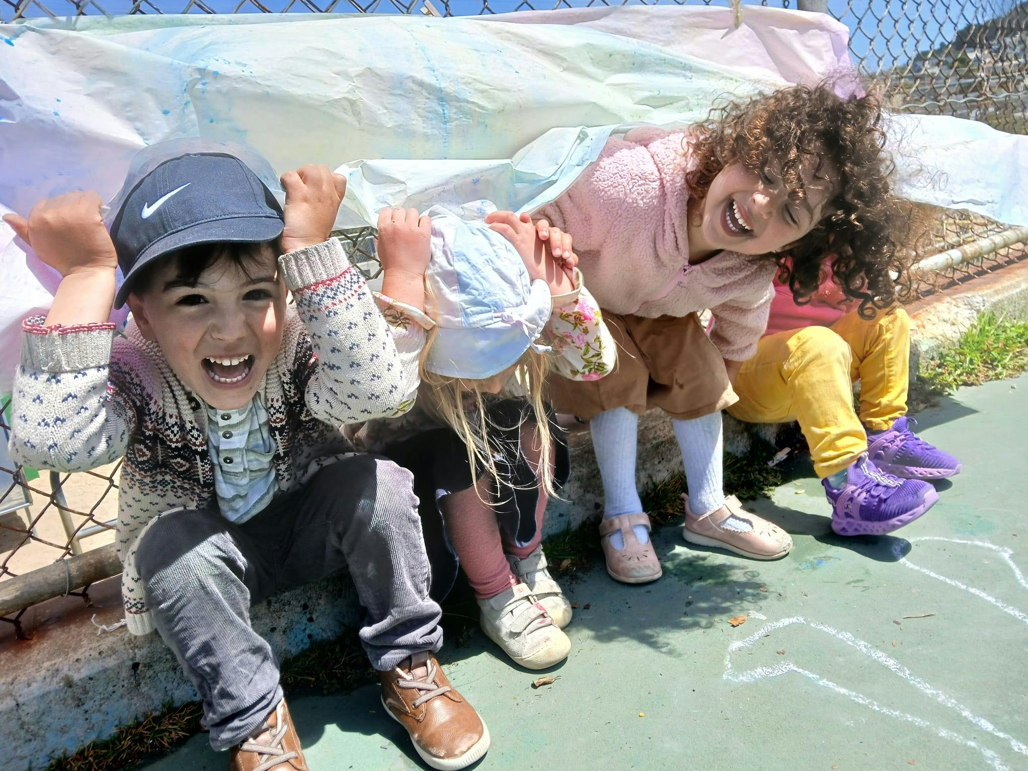 Three children sitting outside with a sheet over their heads