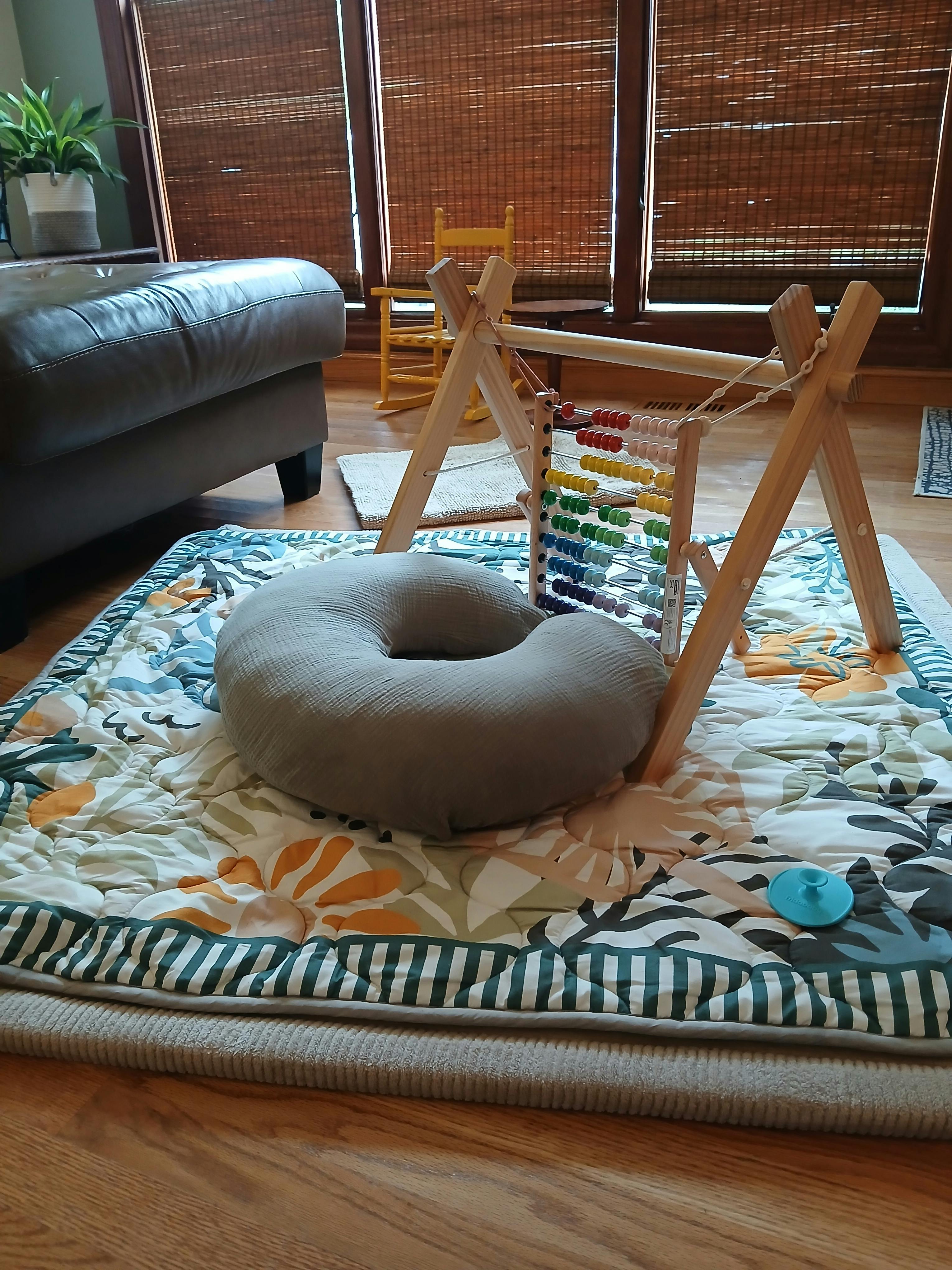 Wooden baby activity gym and a grey nursing pillow on a patterned play mat