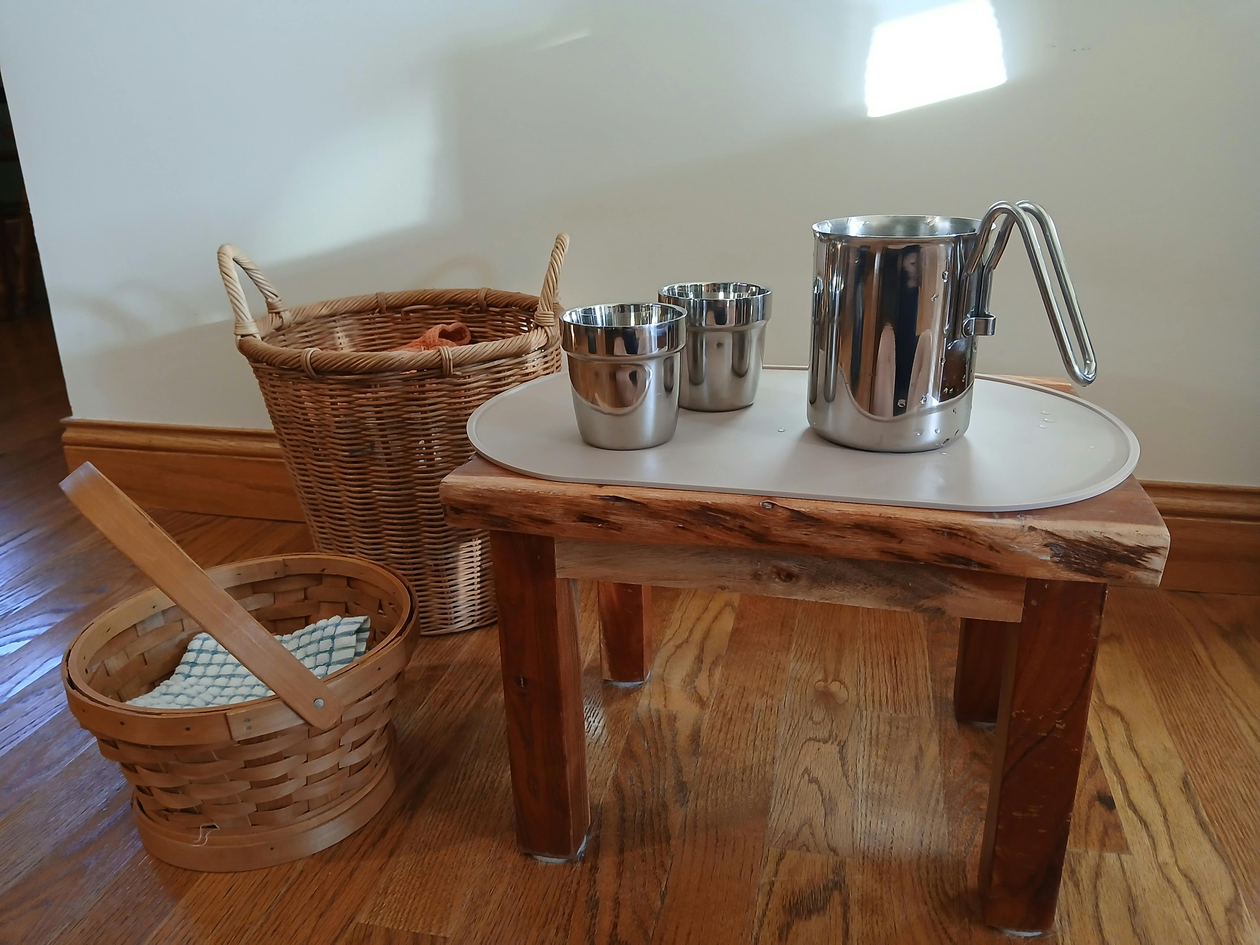 Stainless steel cups and a pitcher on a small wooden table with wicker baskets