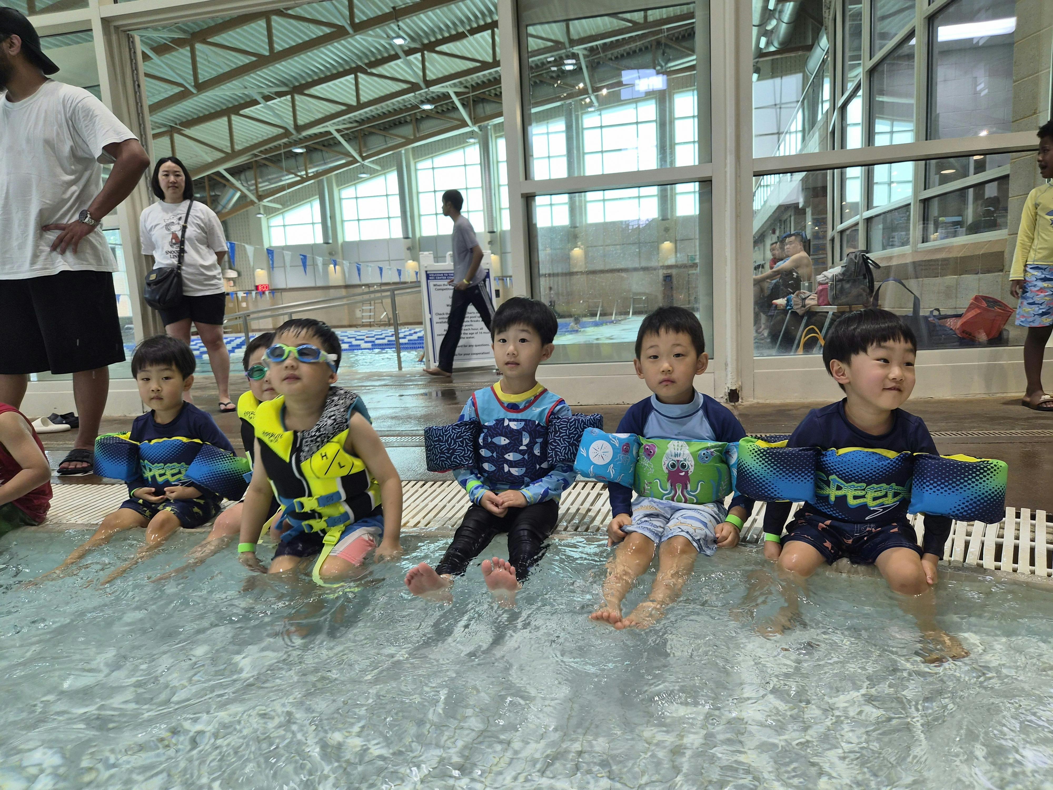 Group of children wearing swim gear and flotation devices sitting by a pool