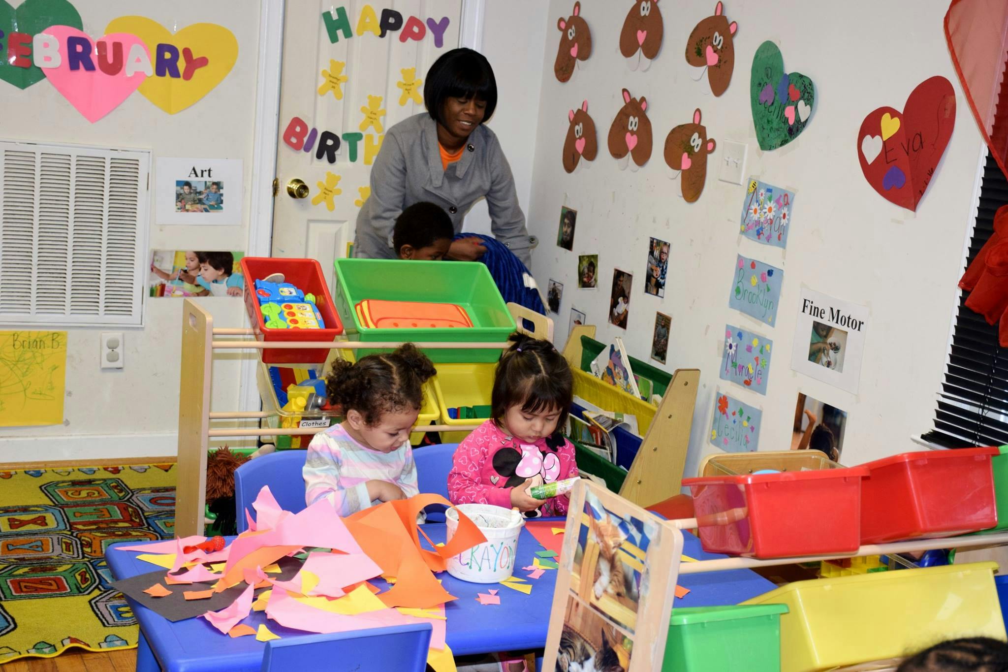 Two children doing crafts at a table with an adult supervising in the background