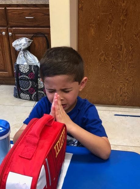 Child with hands pressed together, eyes closed, next to a red bag