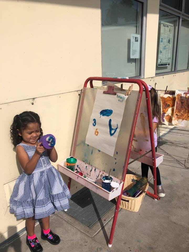 Child holding a purple cup next to an easel with a painting