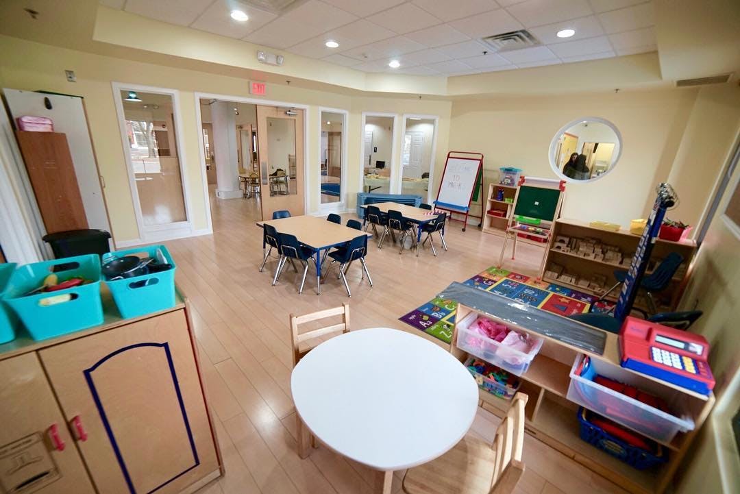 Classroom with small tables and chairs, play rug, and shelves with toys and art supplies