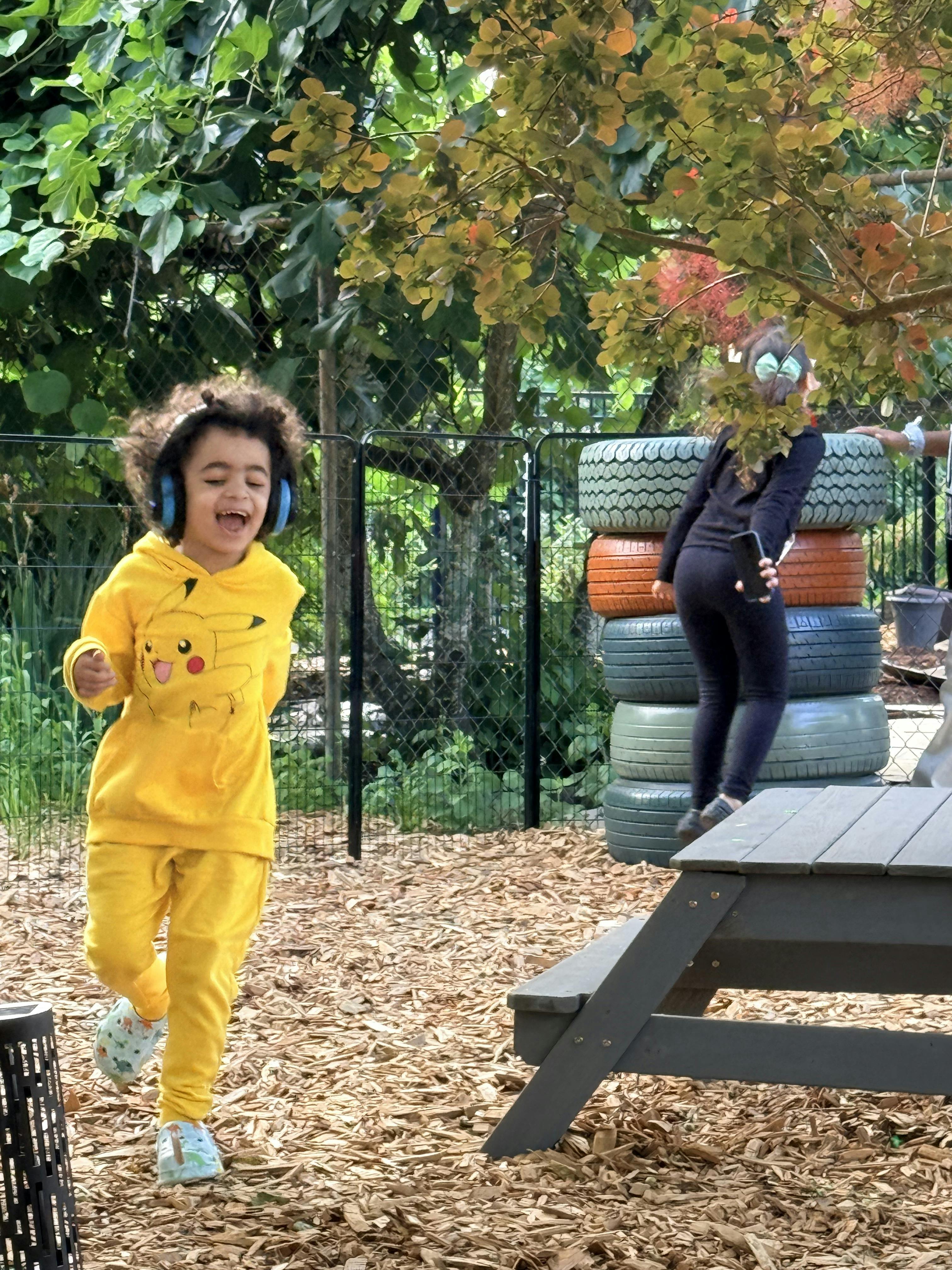 Child wearing yellow Pikachu outfit and blue headphones, another child climbs on tires