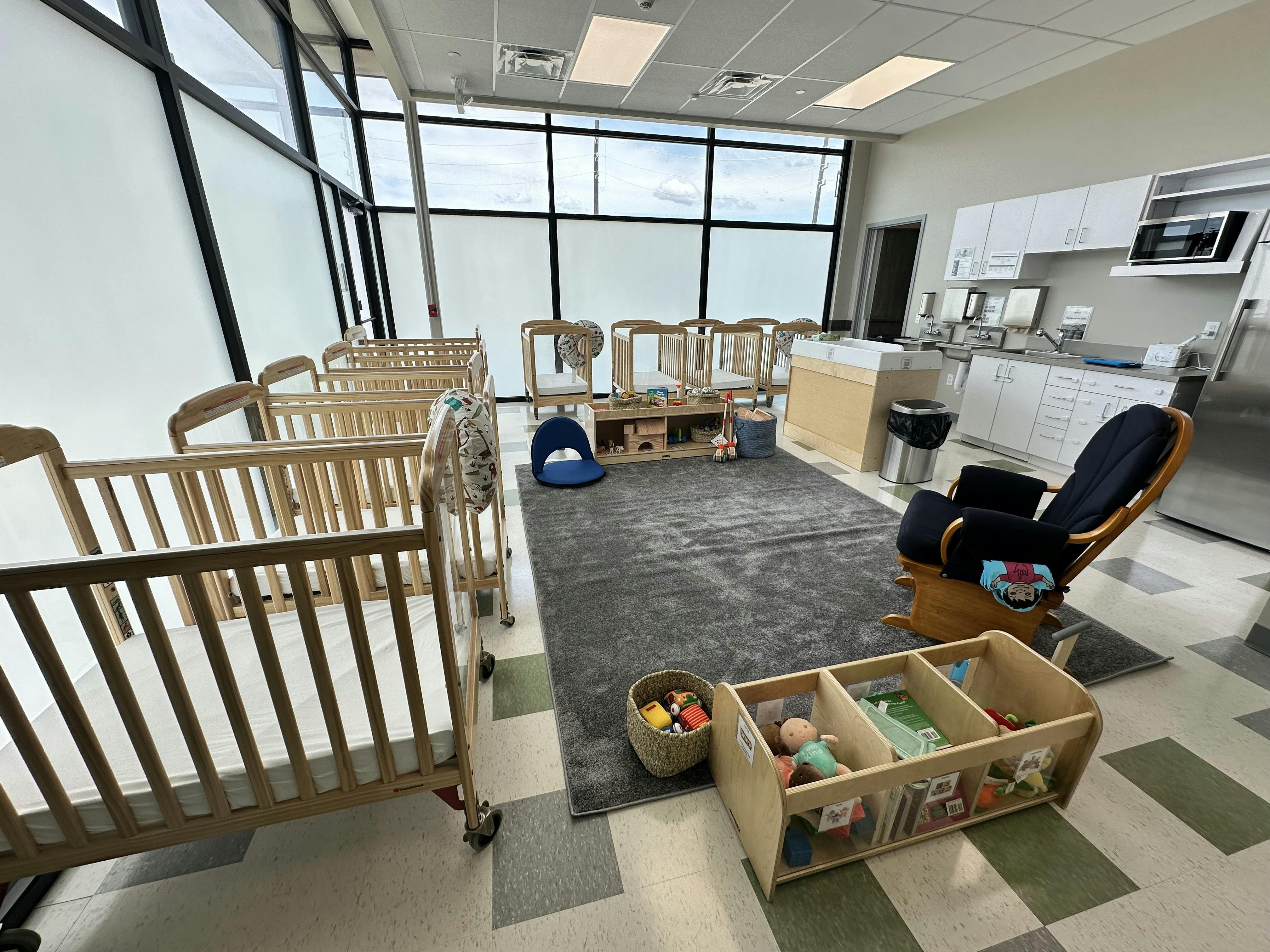 Room with multiple cribs, a rug, a rocking chair, and a play kitchen area