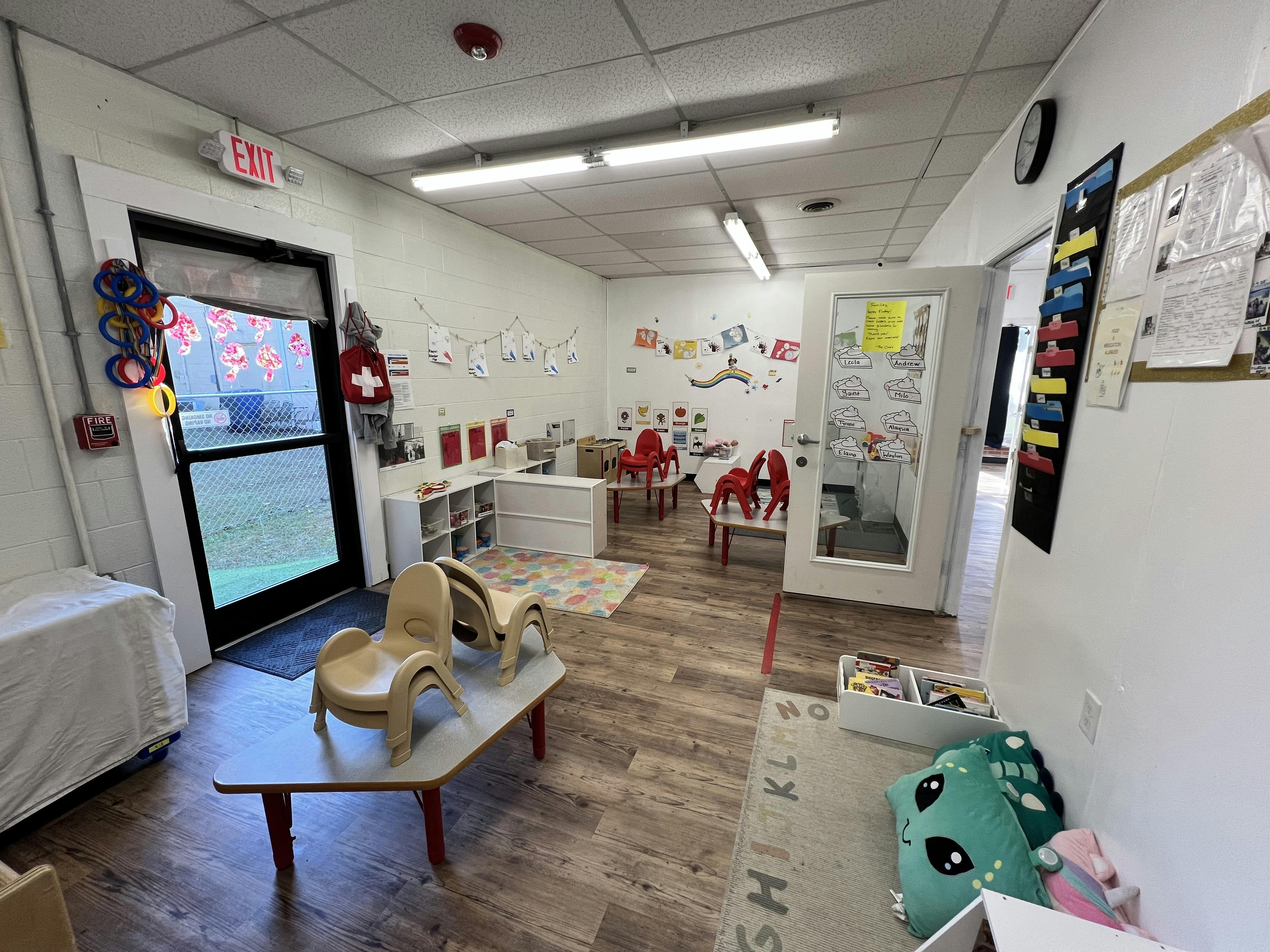 An empty classroom with small tables and chairs, shelves, and a door leading outside