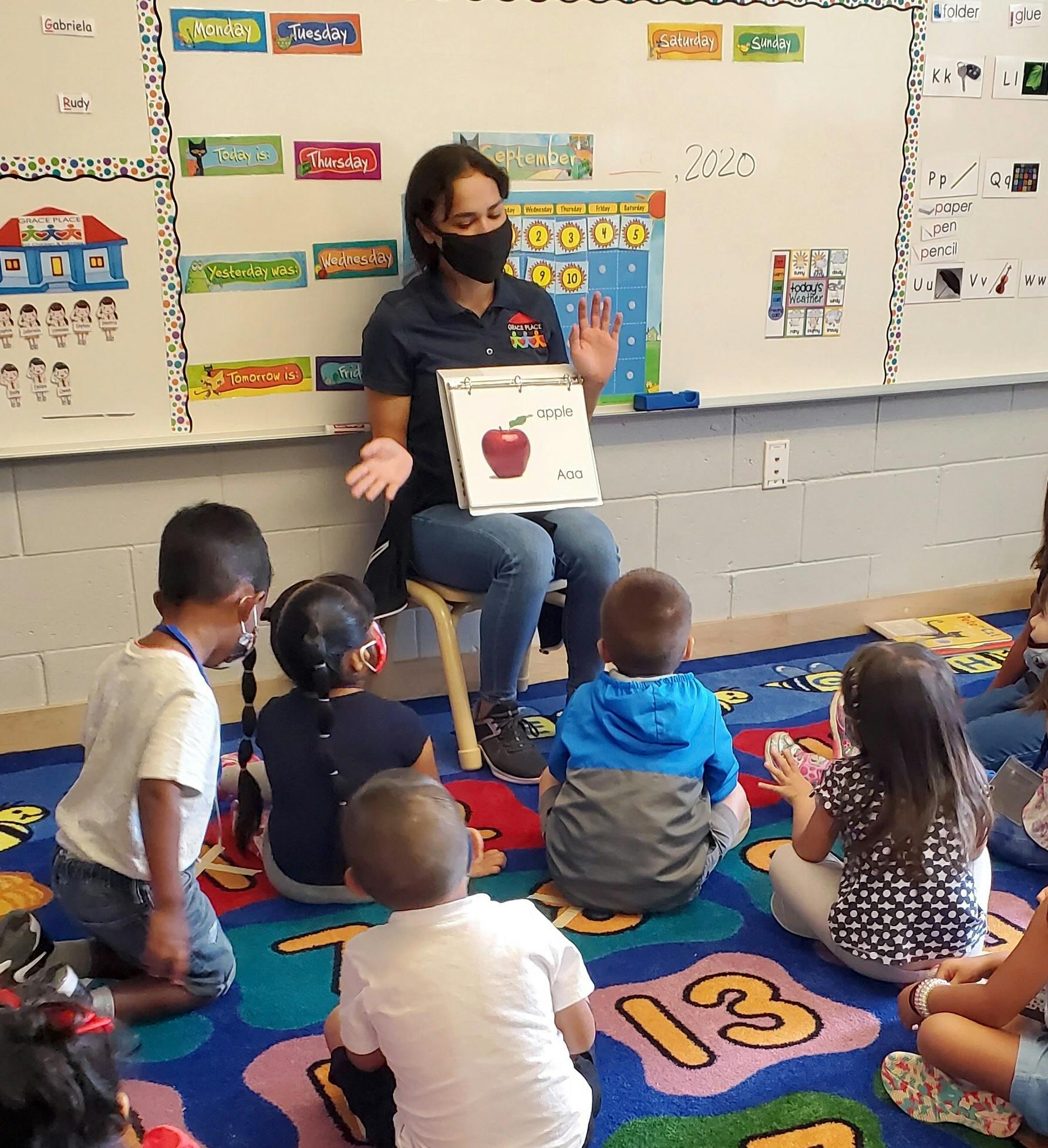 Person in mask teaching children in a classroom, holding up a picture of an apple