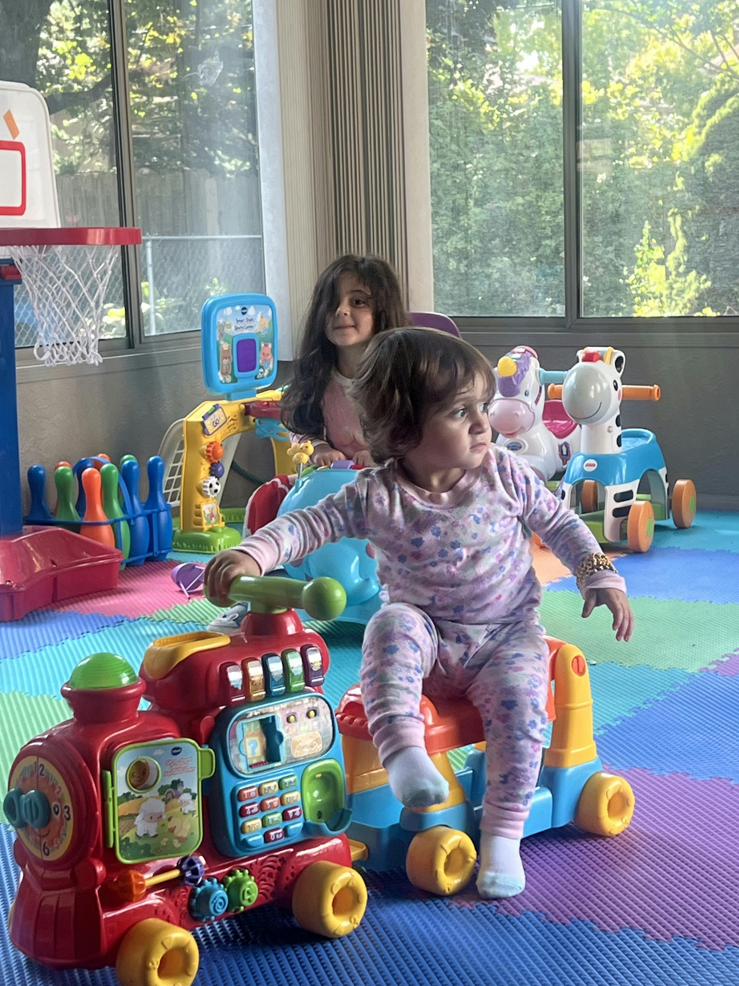 Child riding a toy train with another child smiling in the background
