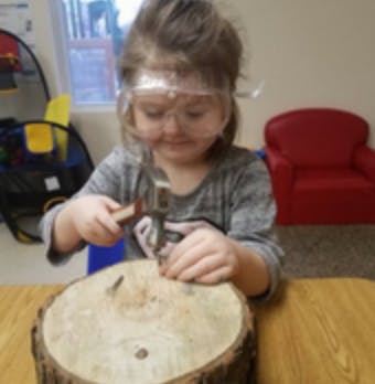 Child wearing safety glasses hammering a nail into a log