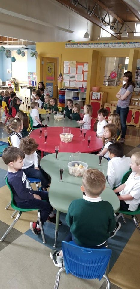 Children sitting at tables in a classroom with an adult standing nearby
