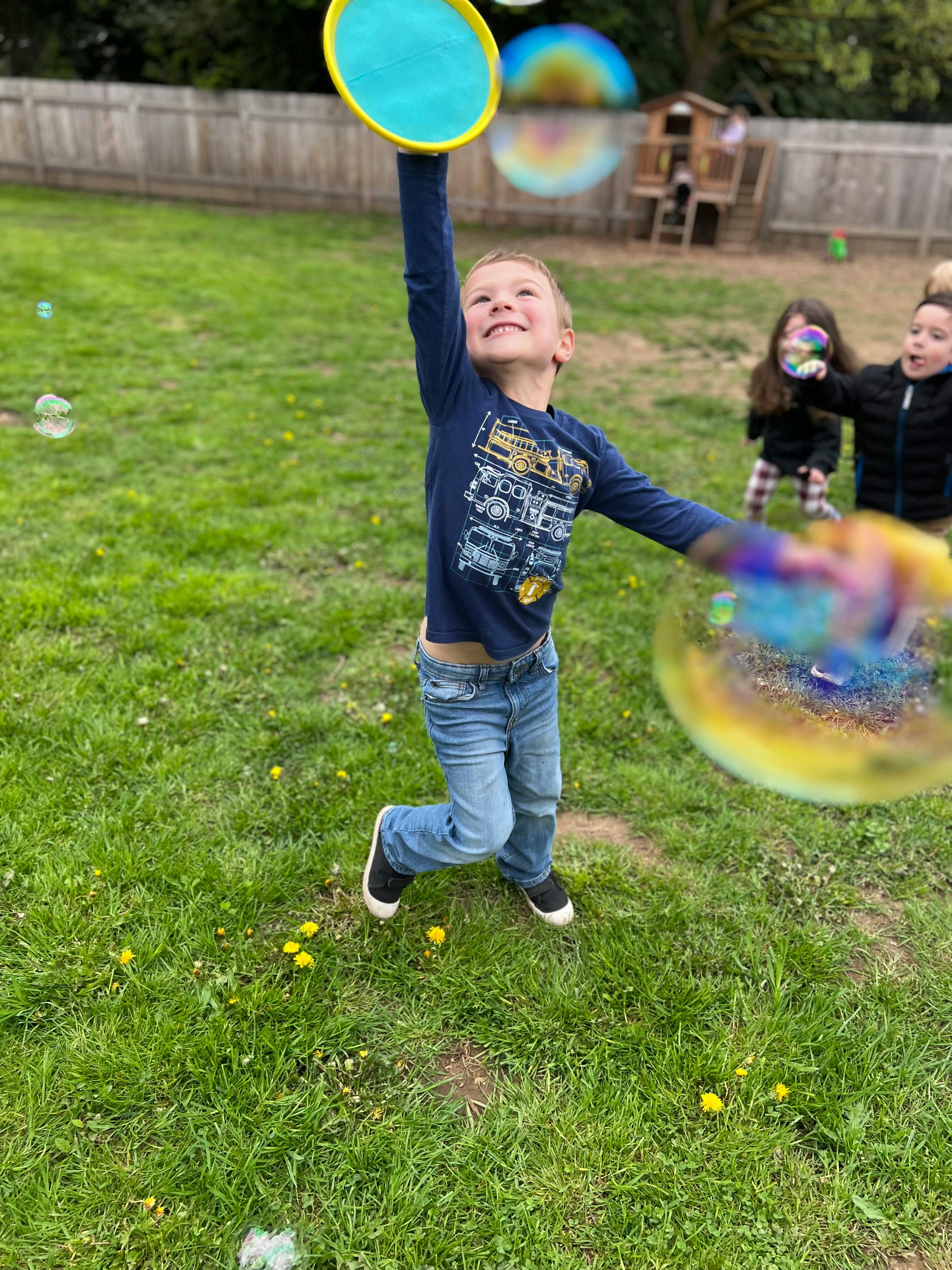 Child reaching up with a paddle to hit bubbles in a grassy yard