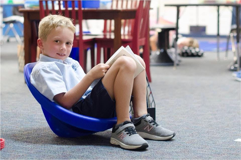 Child sitting in a blue chair and reading a book in a classroom