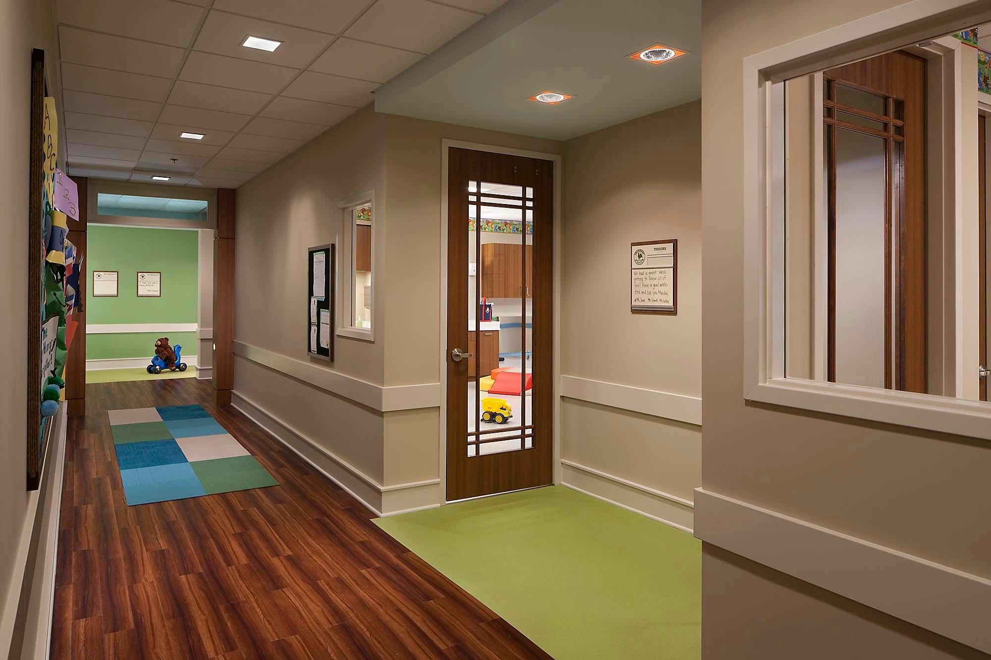 Hallway with wood floor and green carpet leading to rooms with children playing