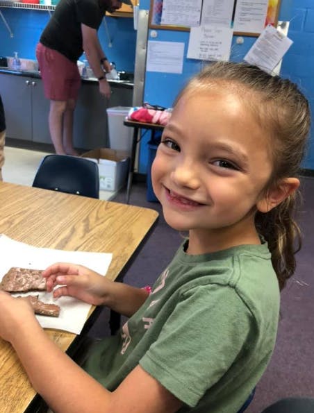 Child at a desk holding a piece of food, smiling at the camera