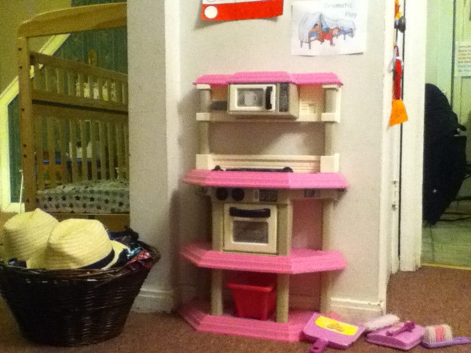 Pink toy kitchen set with a basket of hats and a crib in the background