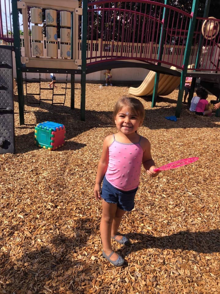 Child standing on wood chips at a playground, holding a pink toy