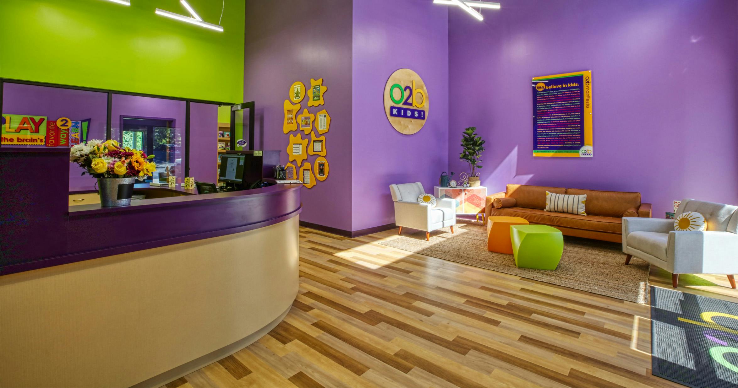 Reception area with a purple desk, a seating area, and a sign that reads "O2B Kids"