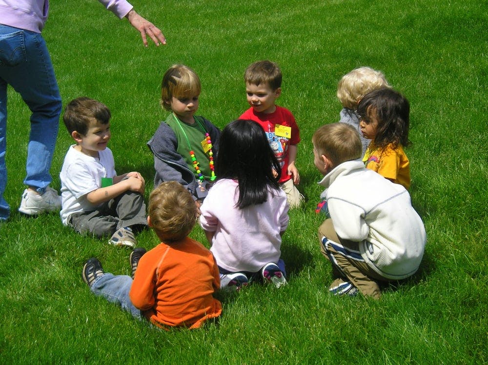 Children sitting in a circle on grass with a person standing nearby