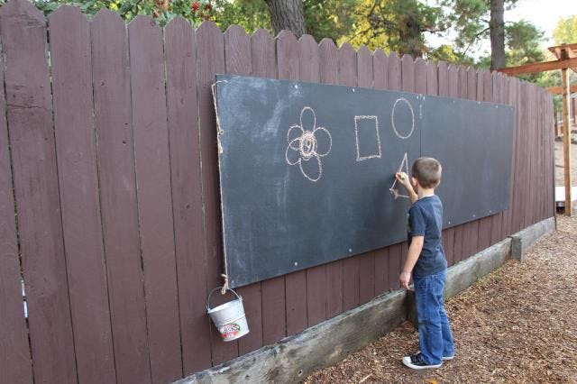 Child drawing on a large chalkboard attached to a wooden fence