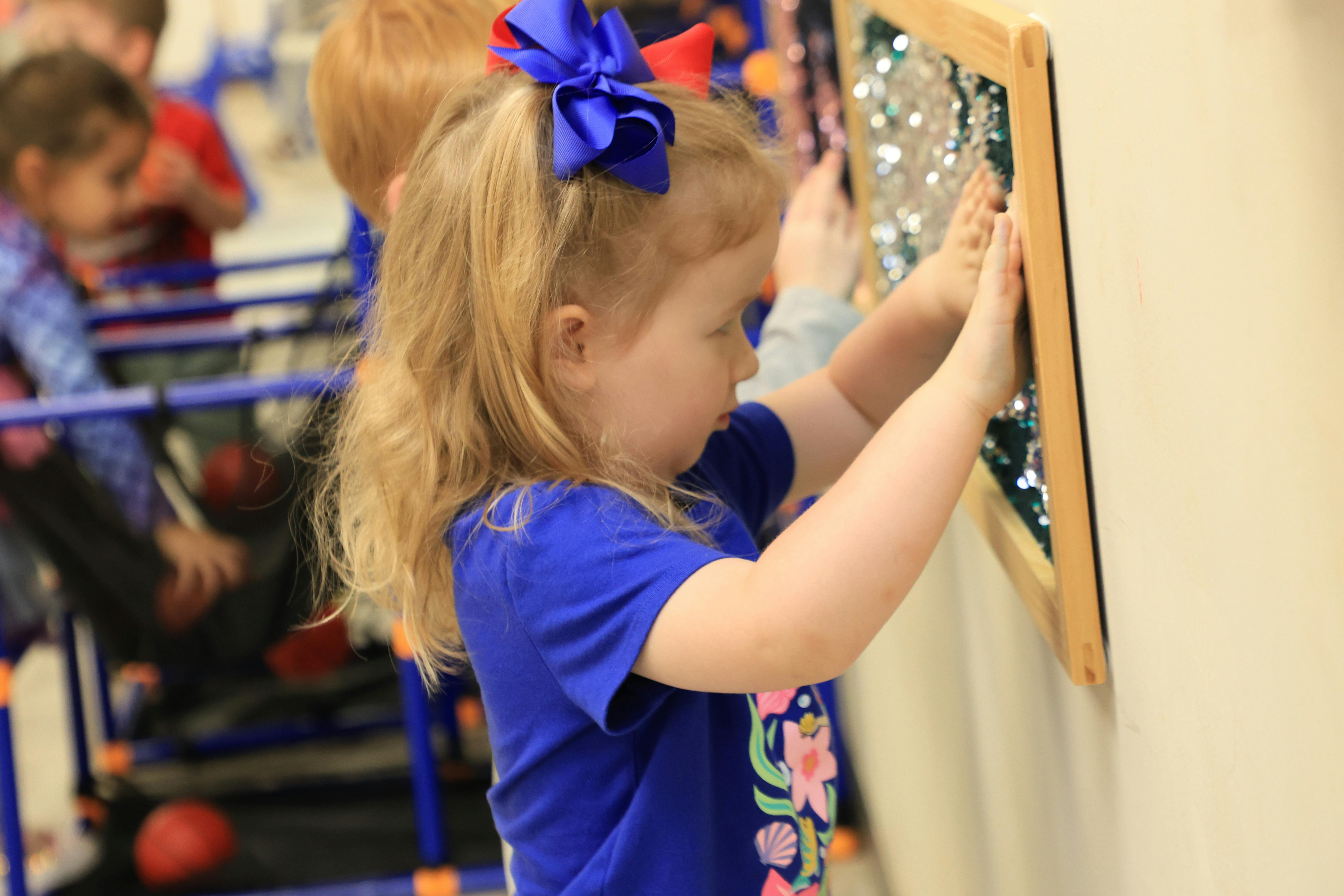 Child with blonde hair and blue bow touching a textured wall panel