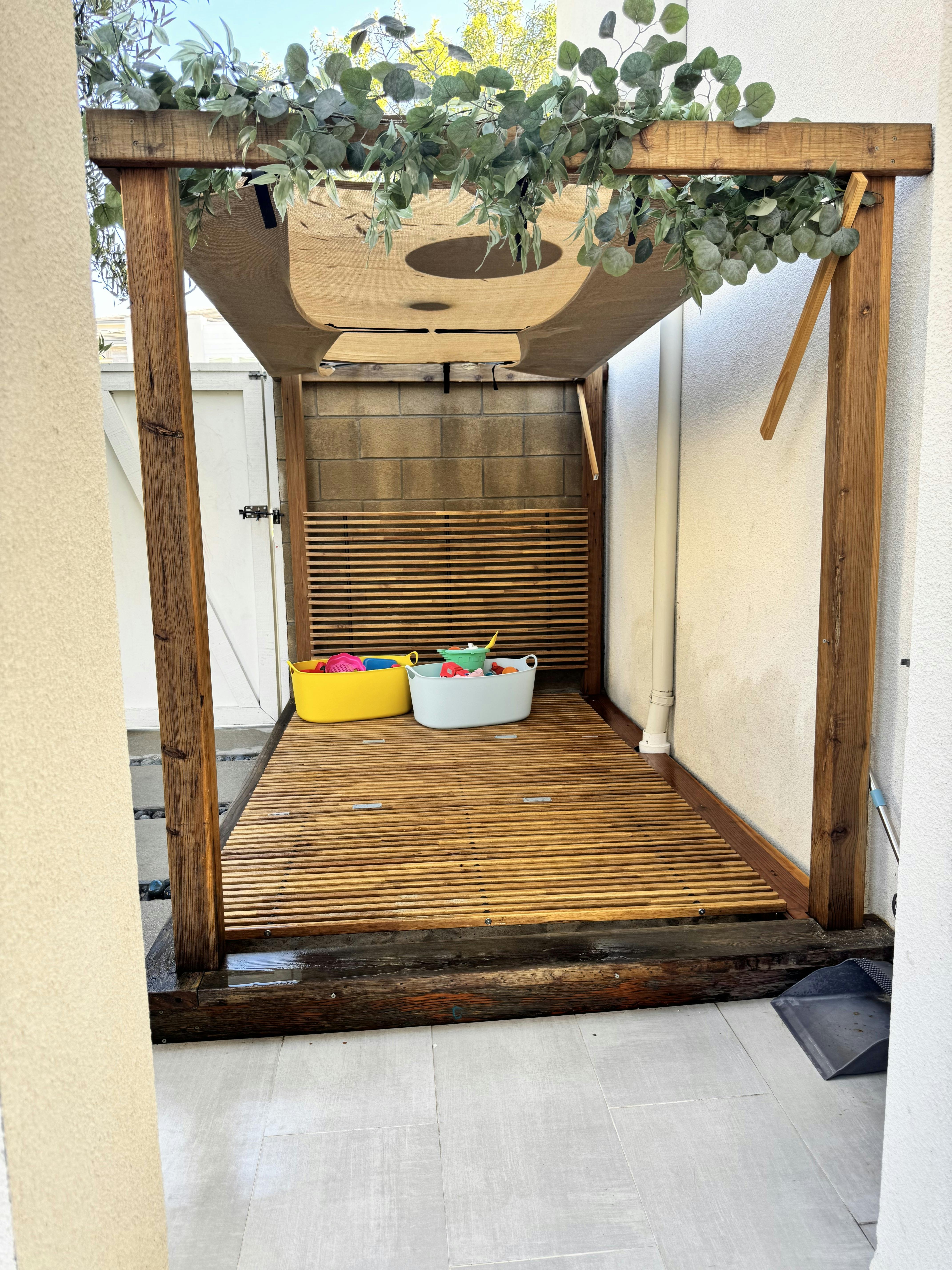 Wooden cabana structure with hanging greenery and toy bins on a slatted floor