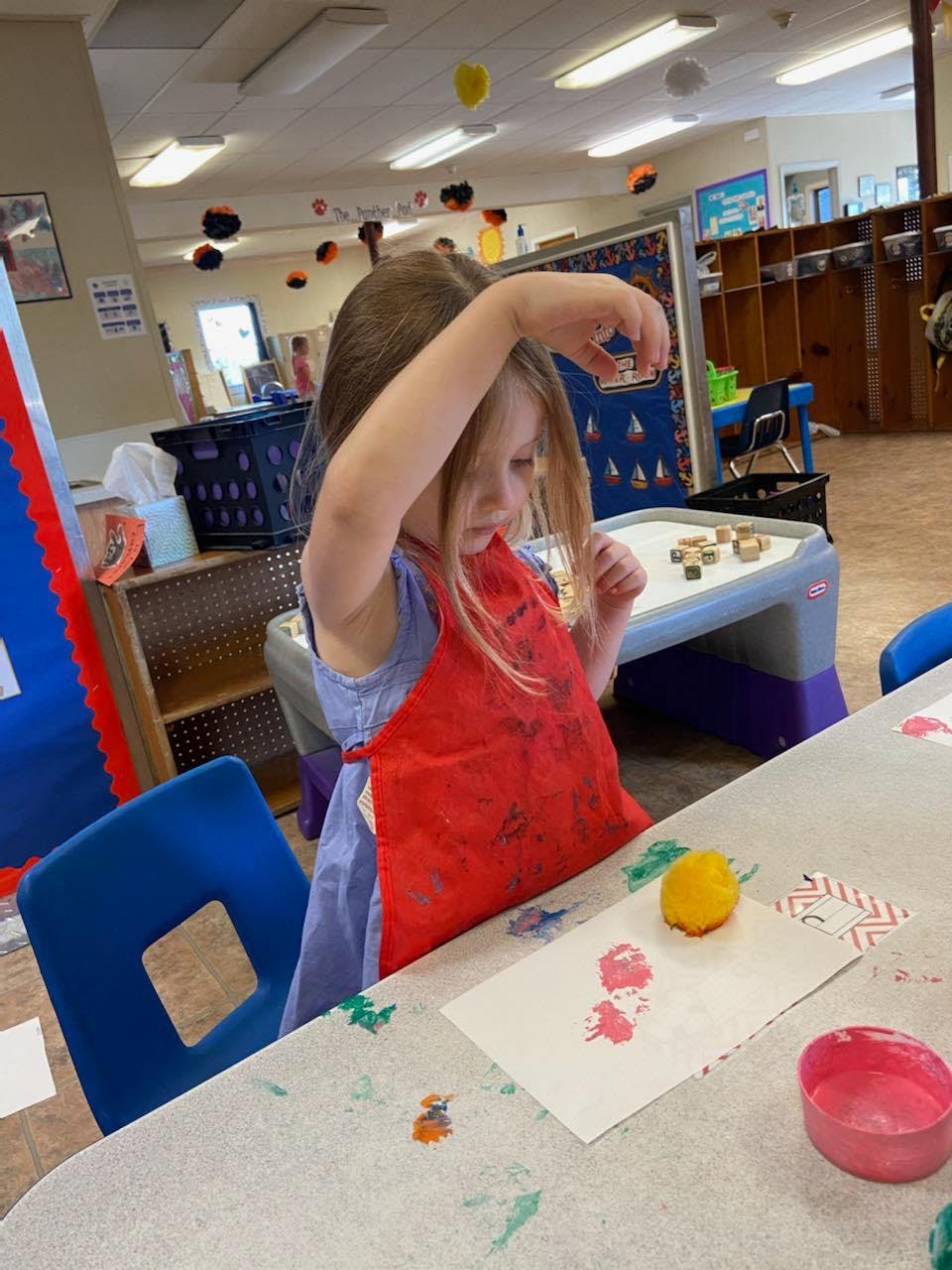 Child in a red apron painting at a table in a classroom