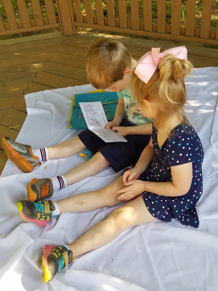 Two children sitting on a blanket, one reading a book