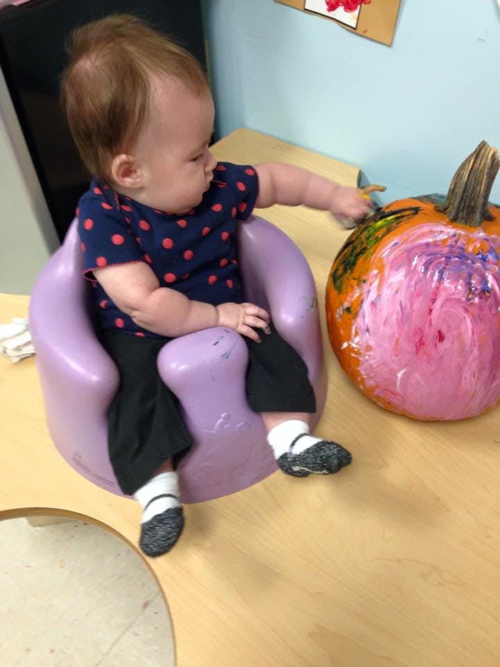 Child in a purple seat reaching for a painted pumpkin