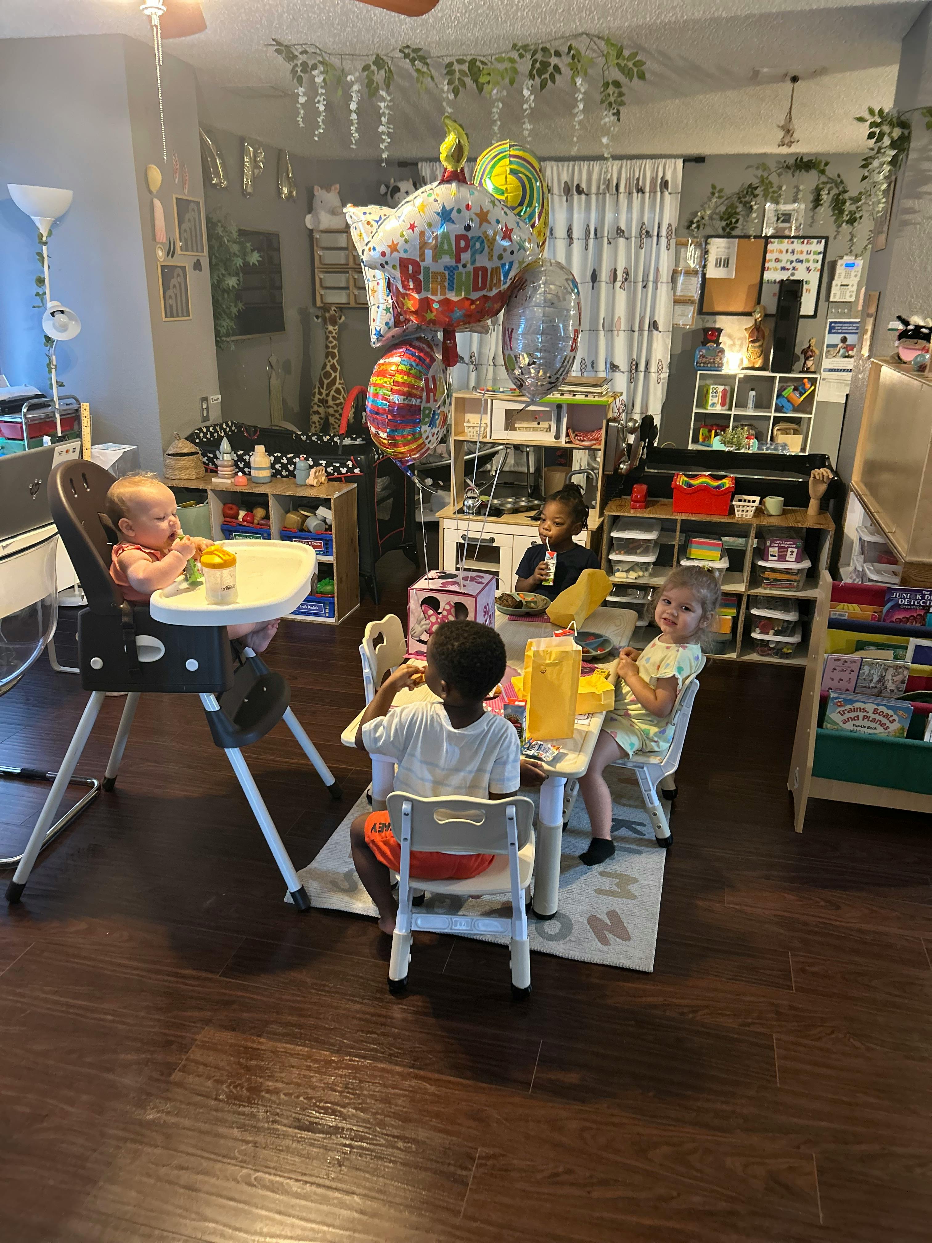 Four children sitting at tables, one in a high chair, with balloons nearby