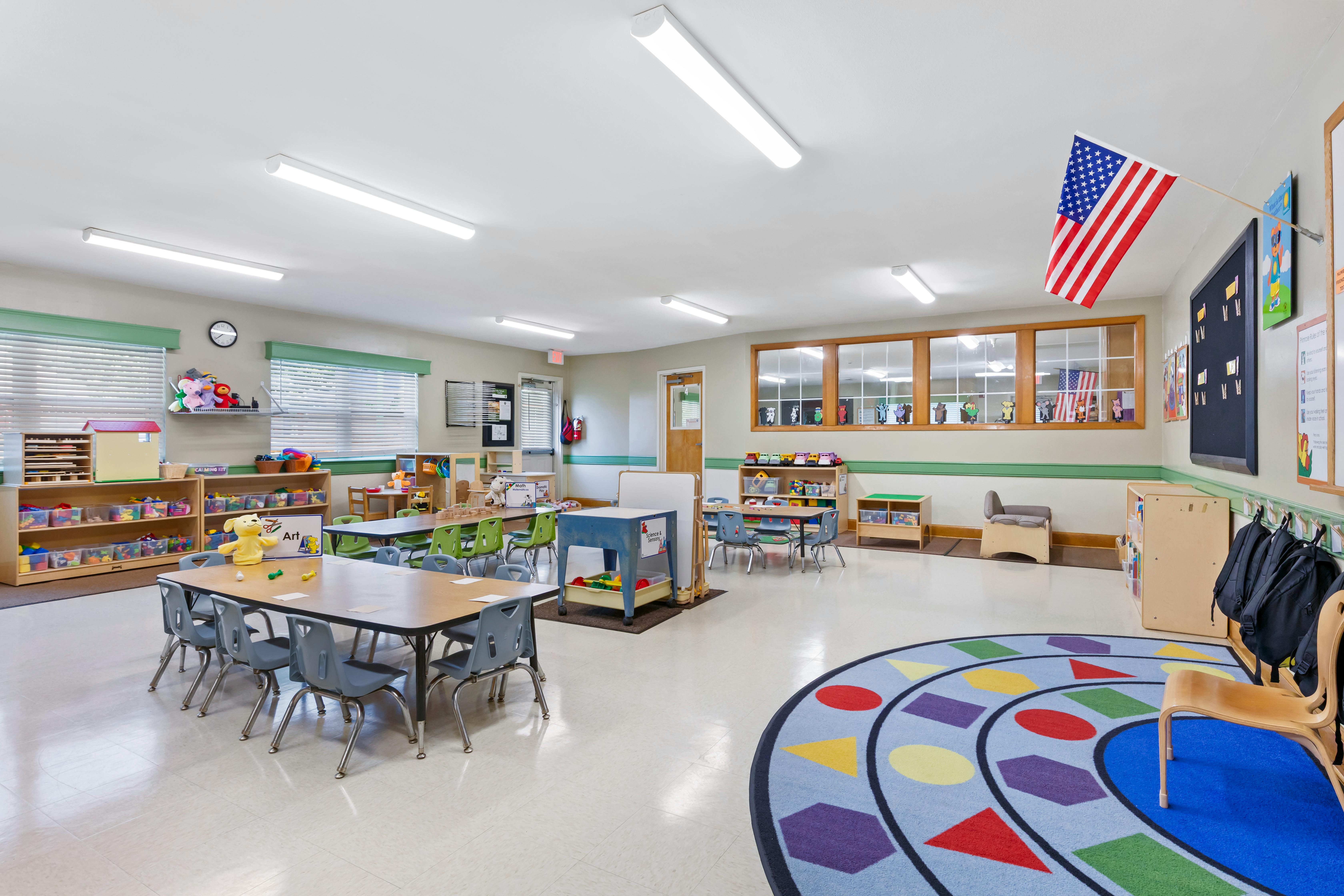An empty classroom with tables, chairs, shelves, and a colorful rug
