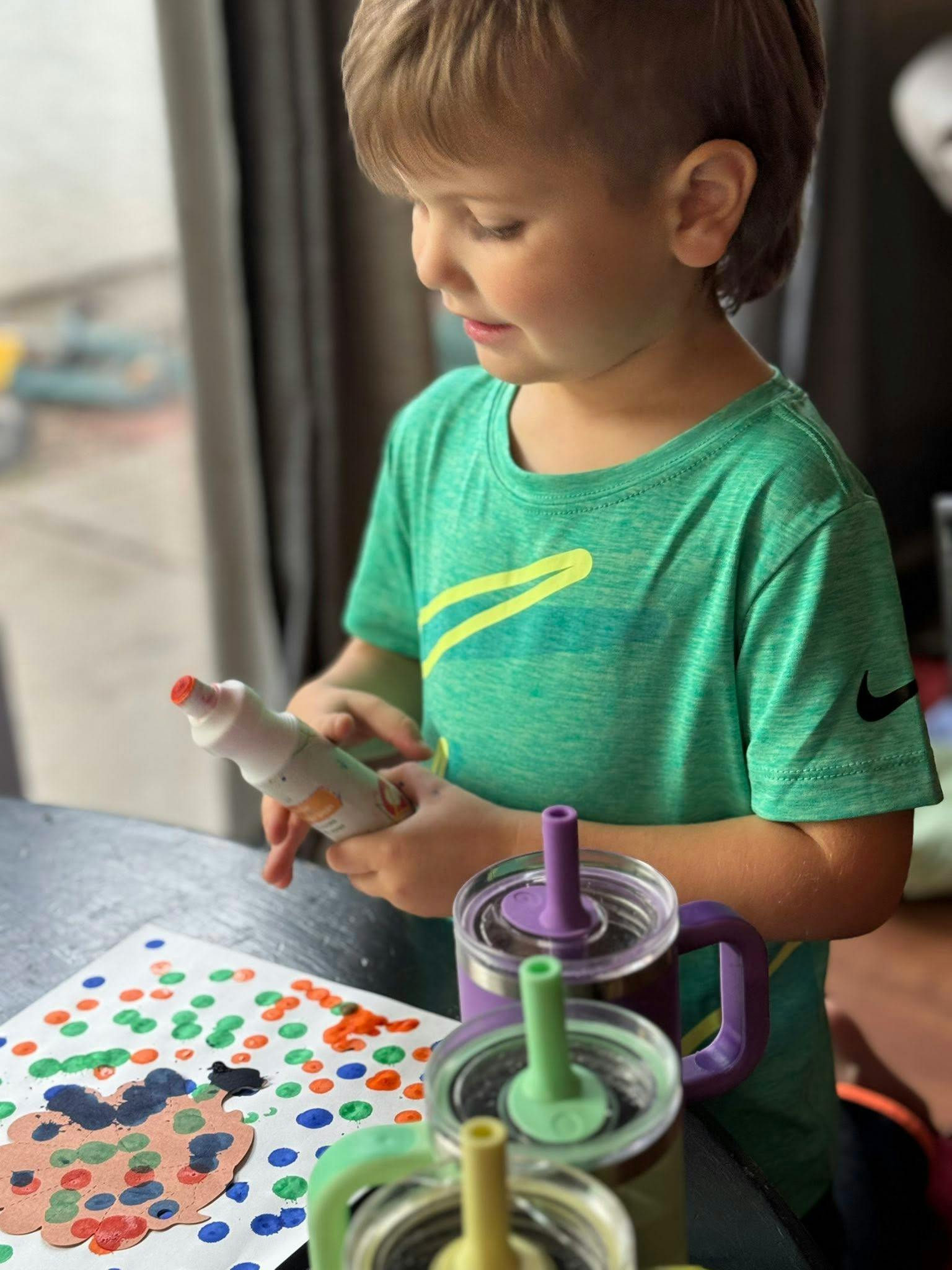 Child holding a paint bottle over a paper with colorful dots