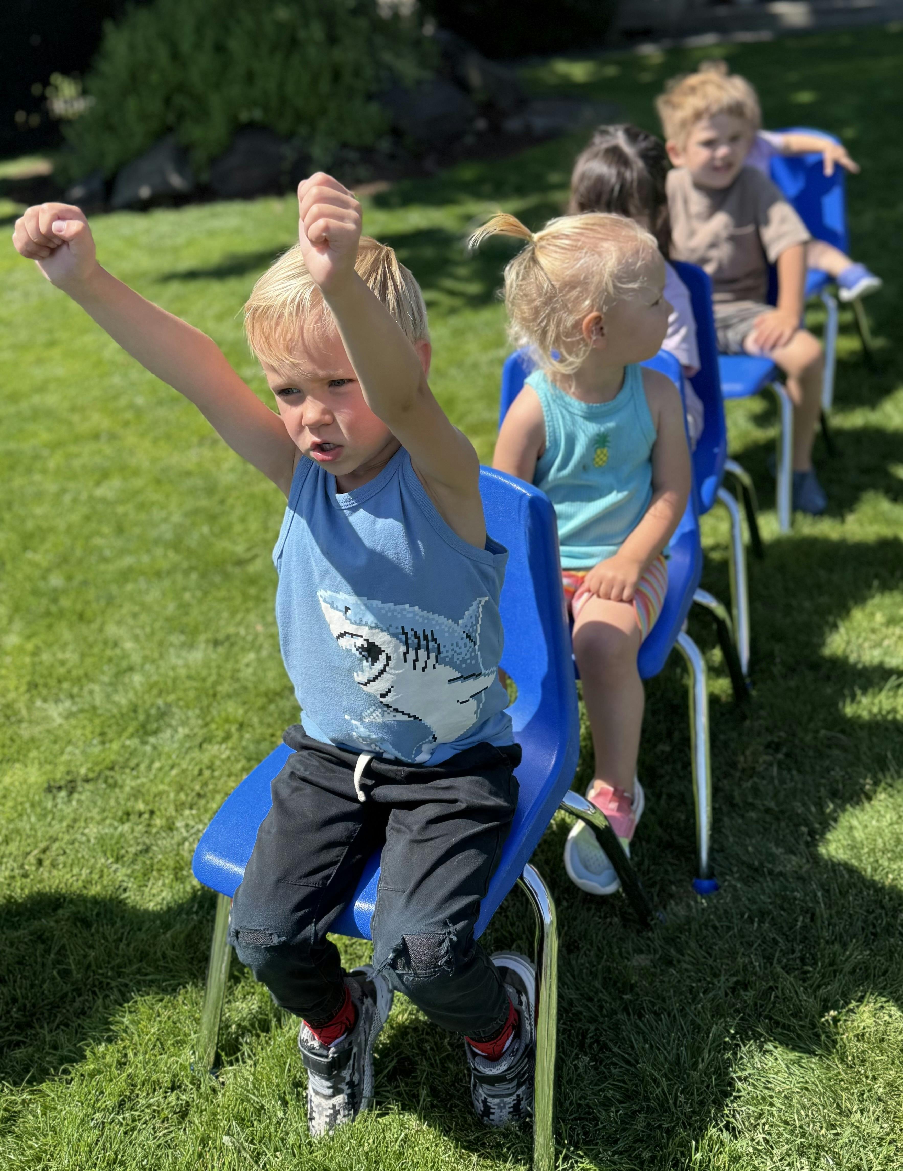 Children sitting in a row of blue chairs on a lawn