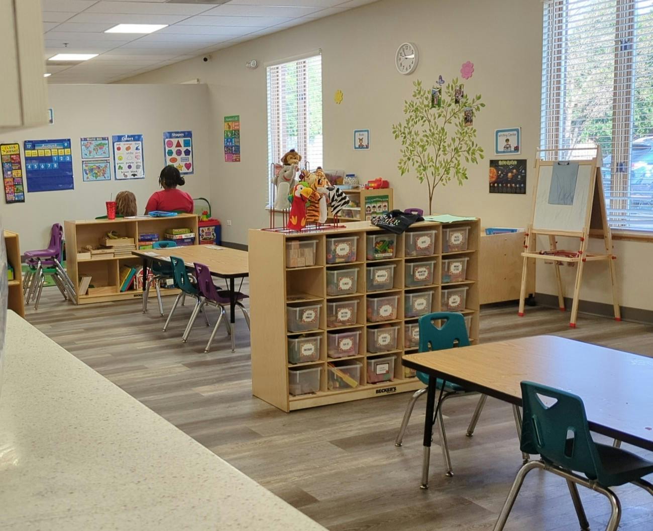 Classroom with tables, chairs, storage units, and a person seated at a desk