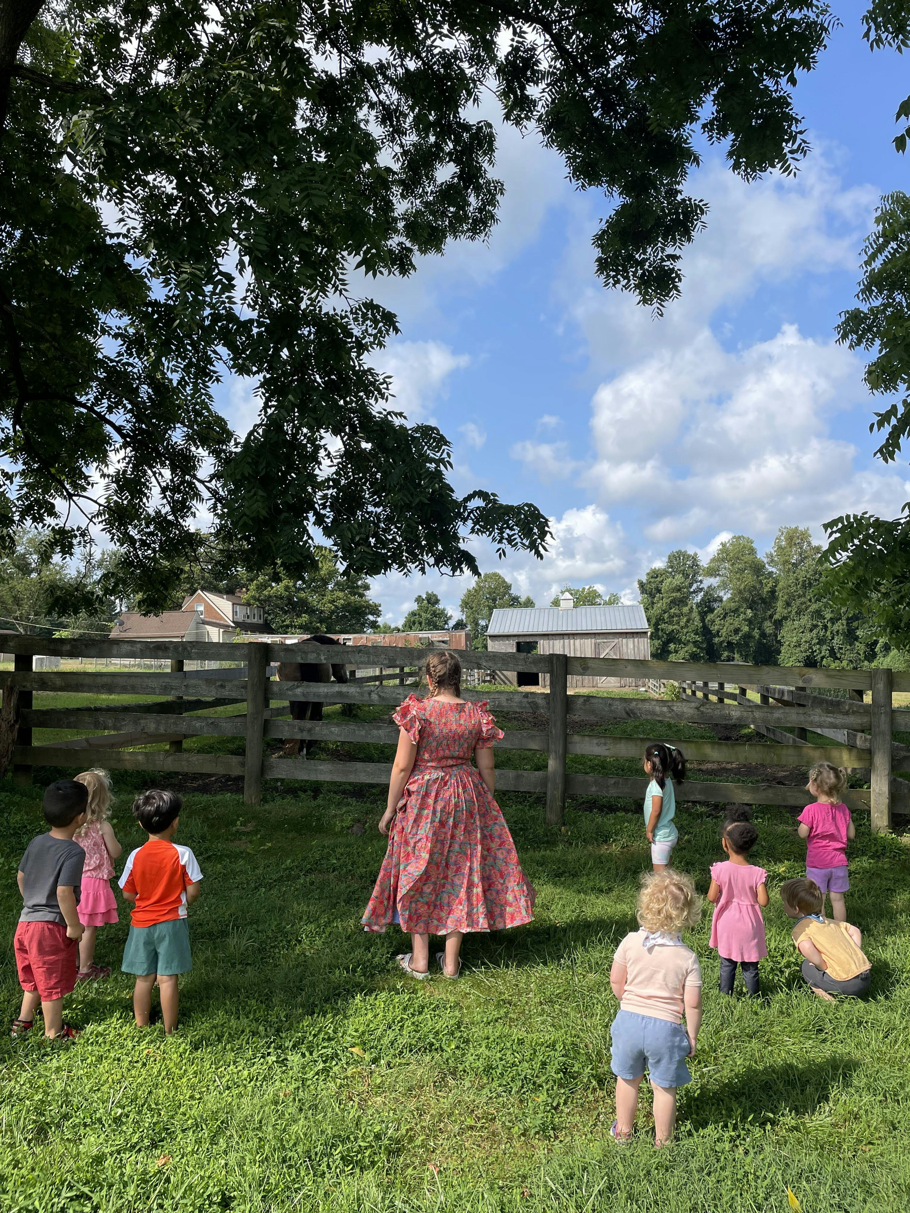 Adult and several children gathered in a grassy area, looking towards a fence and barn