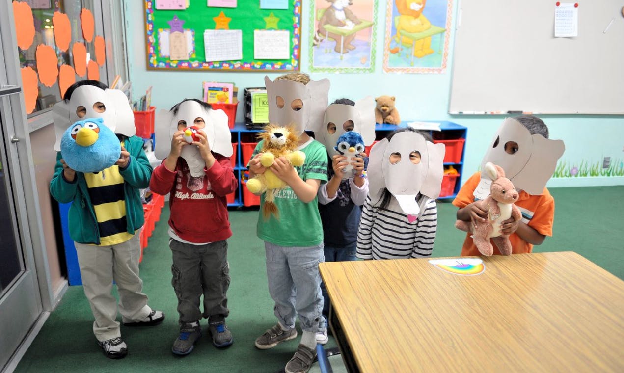 Children wearing paper masks and holding stuffed animals in a classroom