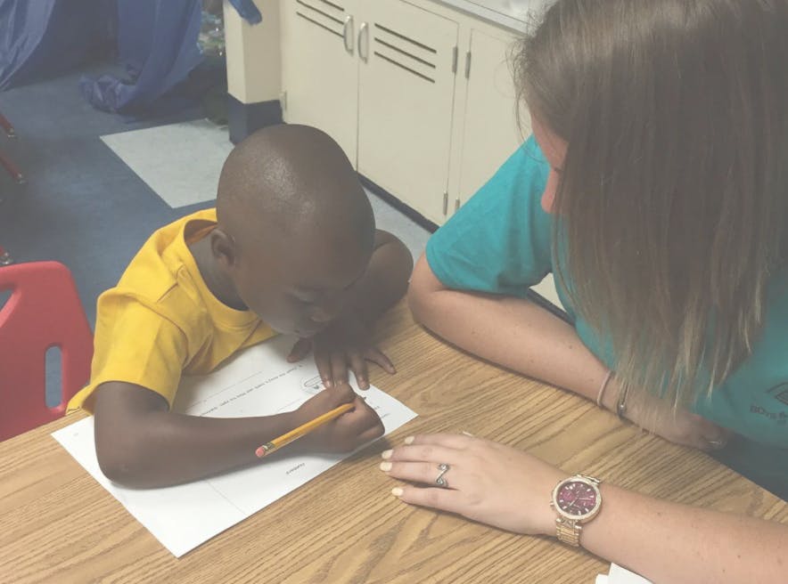 Child writing on paper with a pencil, person observing