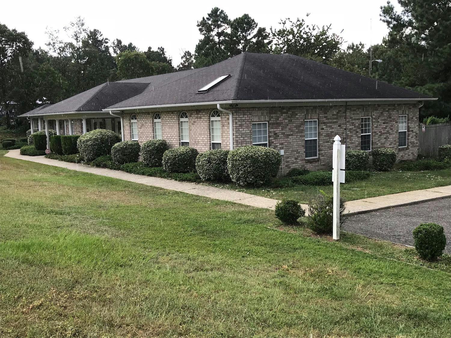 Brick building with a dark roof and bushes along a sidewalk