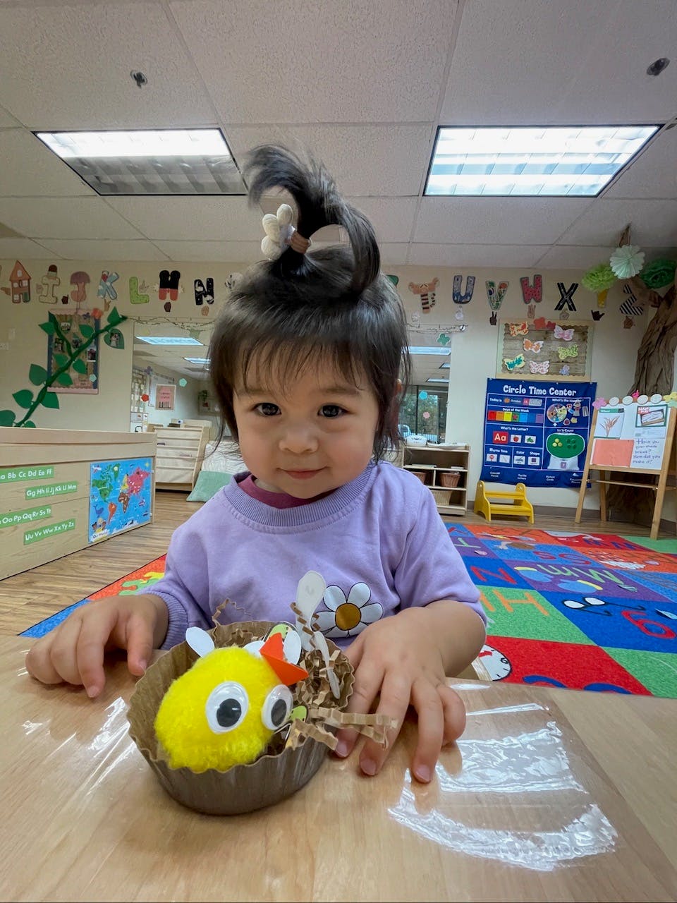 Child with a top bun hairstyle playing with a yellow craft in a classroom