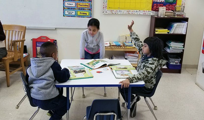 Three children reading books at a table in a classroom, one with a raised hand