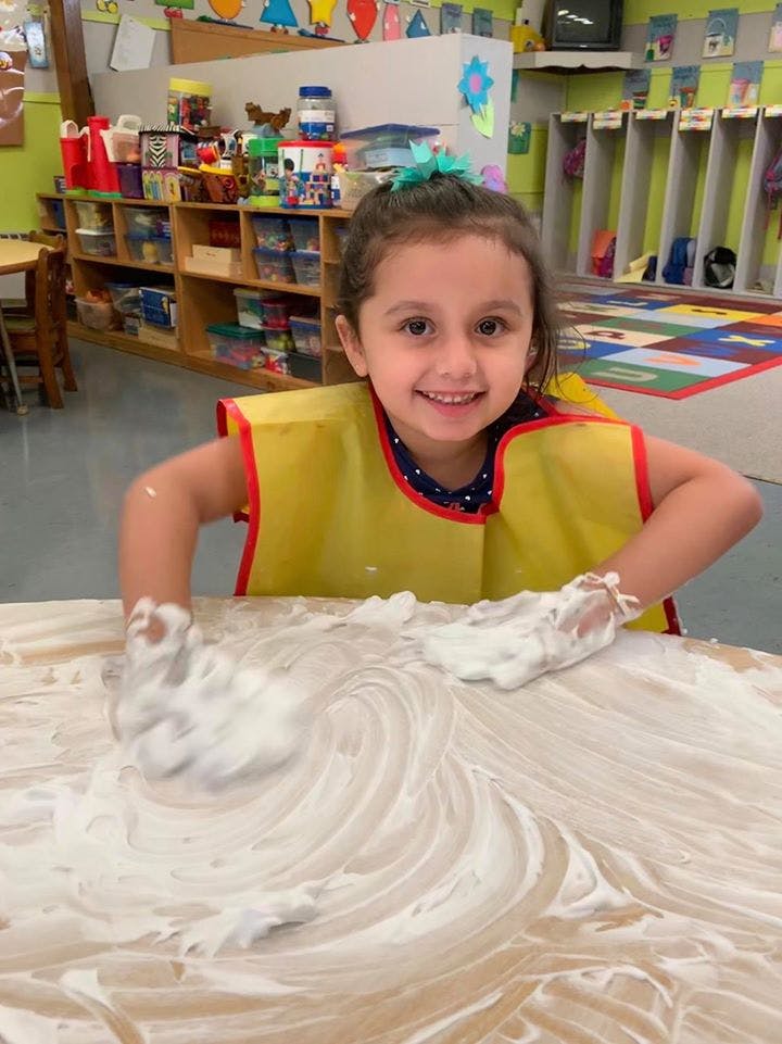 Child in a yellow smock playing with shaving cream on a table in a classroom