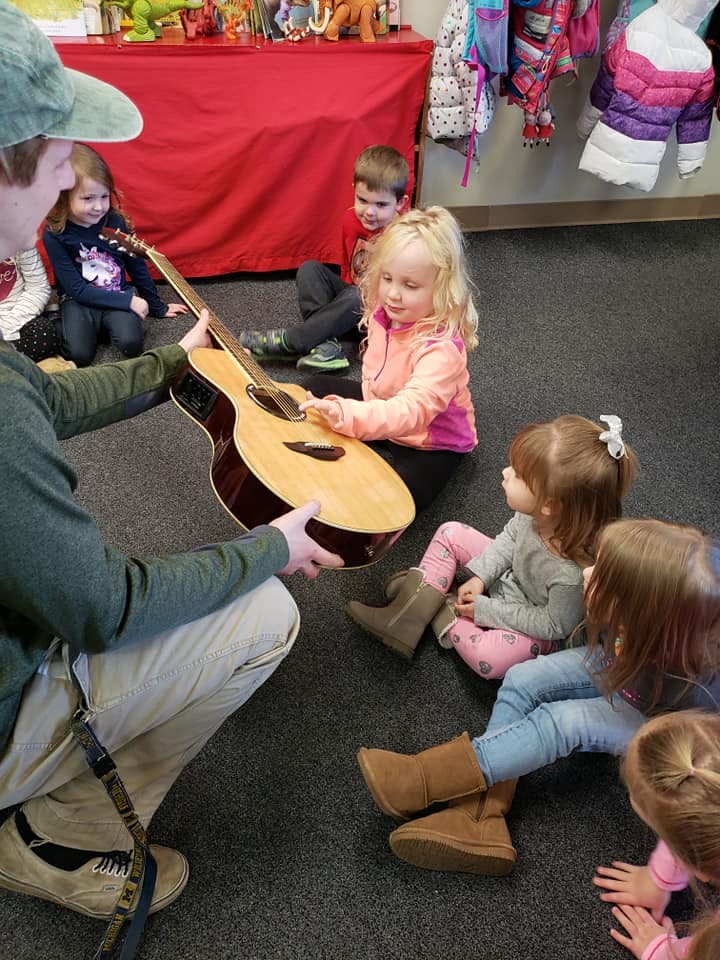 Person holding a guitar for a child to touch, other children watching