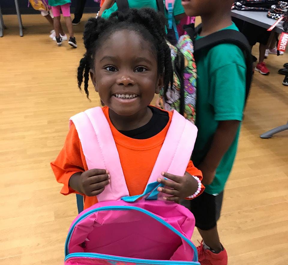Child wearing an orange shirt and pink backpack