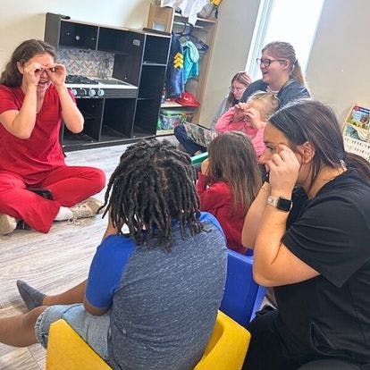 Adult and children sitting on the floor, one adult making a binocular gesture