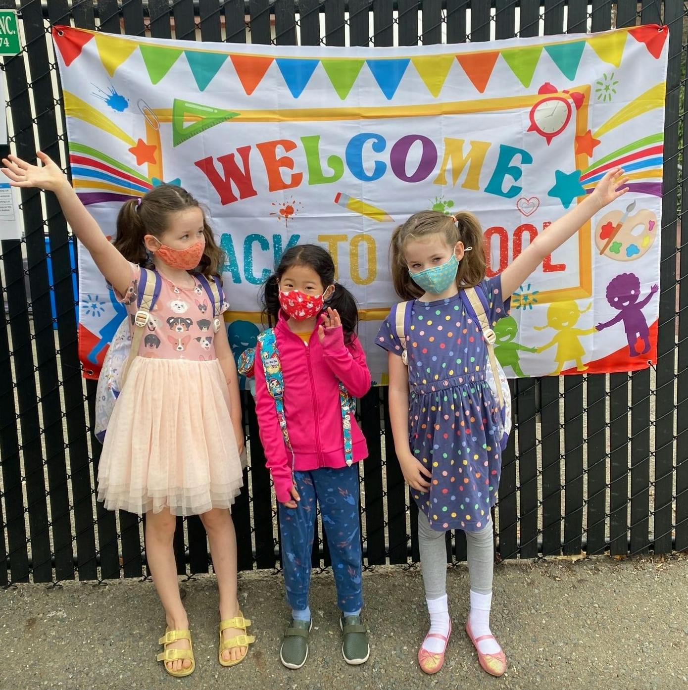 Three children wearing masks and backpacks in front of a "Welcome Back to School" banner